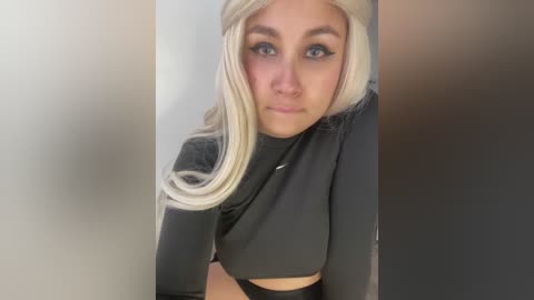 Media: Video of a young woman with long, platinum blonde hair, wearing a black crop top, and black pants, standing against a blurred background.