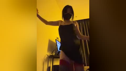 Media: Video of a slim Asian woman in a black dress, black face mask, and red stockings, standing against a yellow wall with a computer screen visible behind her.