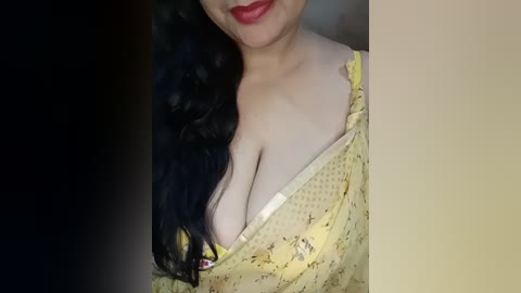 Media: Video of a light-skinned woman with long black hair, wearing a yellow floral-patterned dress with a low neckline revealing ample cleavage. She has bright red lipstick. The background is a blurred, neutral-toned gradient.