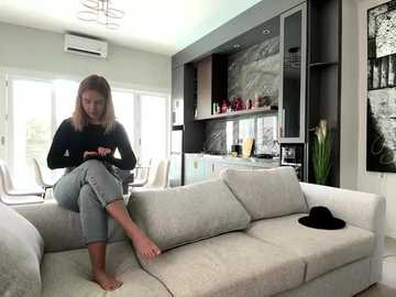 Media: Video of a modern, light-filled living room with a woman in a black sweater and jeans sitting barefoot on a beige sofa, facing a dark wooden kitchen with glass cabinets, red accents, and a black hat on the sofa.