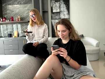 Media: Video of two young women in a modern, minimalist living room. One sits on a sofa, texting, while the other stands, holding a cup, in a chic kitchen.