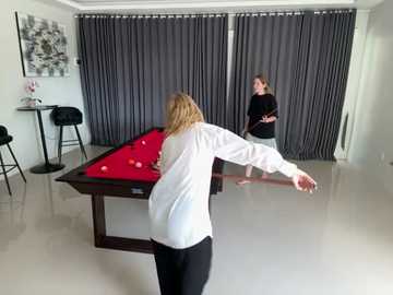 Media: Video of a woman with blonde hair, wearing a white sweater and black pants, playing pool in a modern, minimalist room with grey curtains, white walls, and a red pool table.