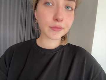 Media: A close-up video of a young woman with fair skin, blue eyes, and light brown hair, wearing a black t-shirt and hoop earrings, with a neutral expression. Background includes a gray curtain and white wall.