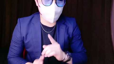 Media: Video of a person in a blue suit, white mask, and round glasses, pointing with both hands, against a dark background.