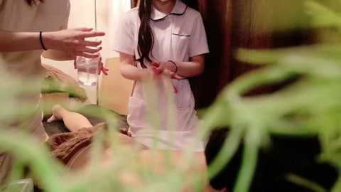 Media: Video of a young Asian girl with long black hair, wearing a white polo shirt, seated on a bed amidst blurred green foliage, holding a glass.