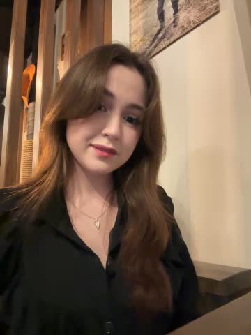 Media: Video of a young woman with fair skin, long brown hair, wearing a black blouse and necklace, indoors with wooden furniture and a framed painting in the background.