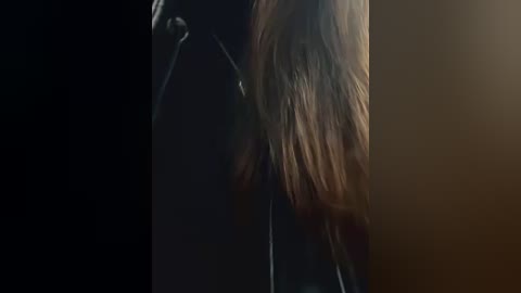 Media: A dimly-lit video of a woman's long, wavy brown hair cascading down her back. She is wearing a black jacket, with her head slightly turned, creating a moody, intimate atmosphere.