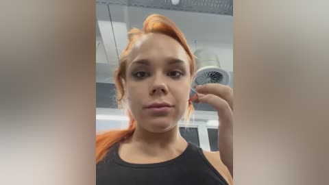 Media: Video of a young woman with light skin and orange hair, wearing a black tank top, taking a selfie in a modern, well-lit bathroom with a white fan and recessed lighting.