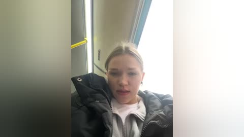 Media: Video of a young blonde woman with fair skin wearing a black puffer jacket and light-colored shirt, sitting inside a dimly lit train compartment, with a yellow handle and window in the background.