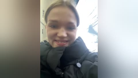 Media: A video of a young, light-skinned woman with blonde hair, smiling broadly. She wears a black, puffy winter jacket with a high collar. The background is blurry, possibly an indoor setting.