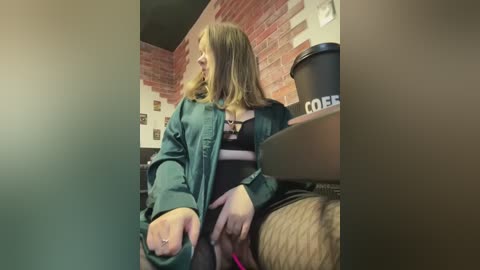 Media: Video of a woman in a teal jacket, sitting on a wooden stool, leaning forward, with a brick wall and coffee cup in the background.