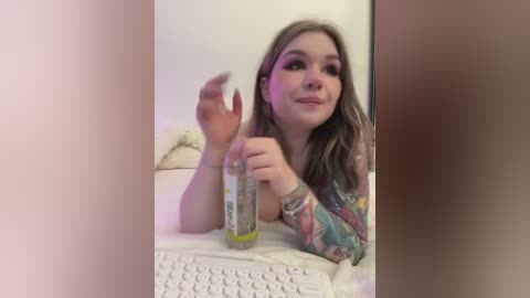 Media: Video of a young woman with long brown hair, wearing a colorful long-sleeve shirt, sitting at a white keyboard. She holds a glass bottle with a yellow lid, smiling and making a playful gesture with her other hand.