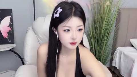 Media: A young Asian woman with long black hair and a pink hair clip, wearing a black halter top, sits indoors, listening to music with white earphones.