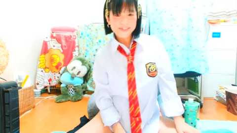 Media: Video of an Asian woman with black hair, wearing a Hogwarts schoolgirl uniform, sitting on a desk in a brightly lit, cluttered room with stuffed animals, toys, and a red tie.