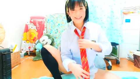 Media: Video of a cheerful Japanese schoolgirl with short black hair, wearing a white shirt, red striped tie, and knee-high black socks, sitting on a floor in a cluttered, brightly lit room with a pink chair and stuffed toys.