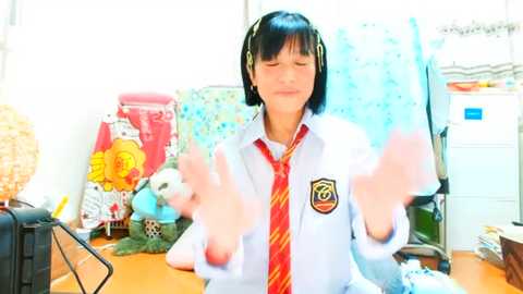 Media: A video of an Asian woman in a white school uniform with a red tie, smiling and making a peace sign in a bright, cluttered office.