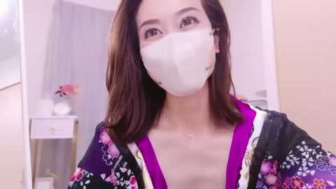 Media: Video of an Asian woman with shoulder-length brown hair, wearing a white face mask, a purple kimono with floral patterns, and standing indoors with a white dresser and a vase in the background.