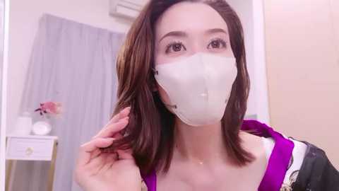 Media: Video of a young Asian woman with fair skin and straight brown hair, wearing a white medical mask and a purple top, standing in a modern bathroom with grey curtains, a white sink, and a pink flower vase.