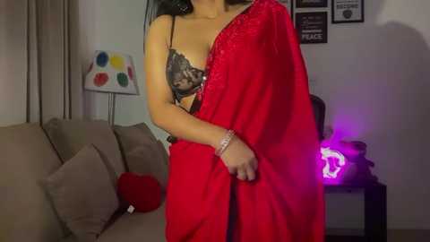 Media: Video of a woman in a black lace bra and vibrant red saree, standing in a modern living room with beige couch, colorful art, and purple LED lights.