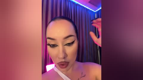 Media: Video of a light-skinned woman with dark hair, wearing dramatic winged eyeliner, red lipstick, and a white strapless dress, standing in a dimly lit room with purple and pink lighting.