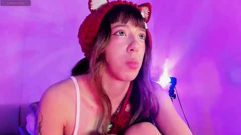 Media: Video of a young woman with fair skin, straight brown hair, wearing a red knit hat with fox ears and a white tank top, sitting in a dimly lit room with purple and blue lighting.