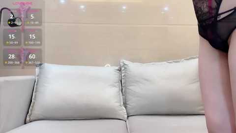 Media: Video of a pale-skinned woman wearing black lace lingerie, standing in front of a white sofa with two light grey pillows. Background shows a white tiled wall with a digital clock and temperature display.