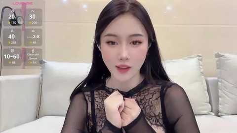 Media: Video of an East Asian woman with long black hair, fair skin, and a slender figure, wearing a sheer black lace top, sitting on a white couch. Background shows a light-colored wall and a digital screen displaying heart rate and other metrics.