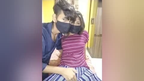 Media: Video of a man with medium skin tone wearing a black mask and blue shirt, hugging a woman with dark skin tone wearing a striped maroon shirt and blue and white striped pants, in a yellow room.