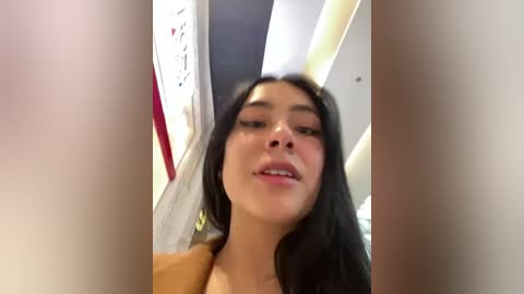 Media: Video of a young woman with long black hair and fair skin, wearing a brown coat, standing in a modern indoor space with white walls and red accents.