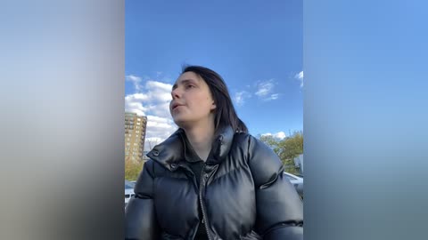 Media: Video of a young woman with long dark hair, wearing a black puffer jacket, gazing upward into a clear blue sky with scattered clouds. Urban background features a high-rise building and trees.