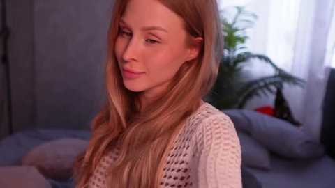 Media: Video of a fair-skinned woman with long, wavy blonde hair, wearing a white knitted sweater, smiling softly. Background features a bed with grey pillows and a green plant, softly lit by natural light.