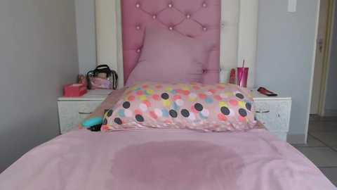 Media: Video of a pink, tufted headboard bed with a pillow featuring a polka-dot pattern in pastel colors. The room has white nightstands with pink accessories, a pink blanket, and a plush toy.