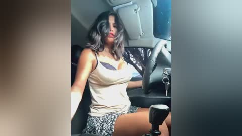 Media: Video of a woman with dark hair, wearing a beige tank top and patterned skirt, sitting in a car, steering wheel in hand.