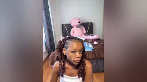 Media: Video of a young Black girl with braided hair and a tear-stained face, sitting on a bed with a stuffed pink bear and messy toys in a cluttered room.