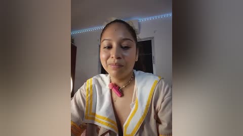 Media: Video of a young woman with light brown skin, wearing a white robe with yellow trim, and a pink necklace, standing indoors with dim lighting and a blue LED strip.