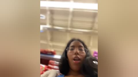 Media: A blurry video of a woman with long black hair, wearing glasses, and a casual outfit, possibly in a grocery store aisle.