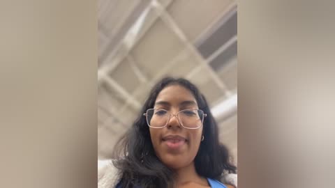 Media: A close-up video of a young woman with medium brown skin, long black hair, and clear glasses, smiling slightly. The background features a blurred, institutional setting with white ceiling panels and fluorescent lighting.