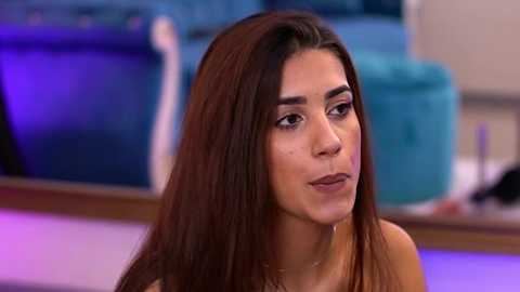 Media: Video of a young woman with long, straight brown hair, light skin, and brown eyes, wearing a brown top, sitting indoors with a teal sofa and purple lighting in the background.