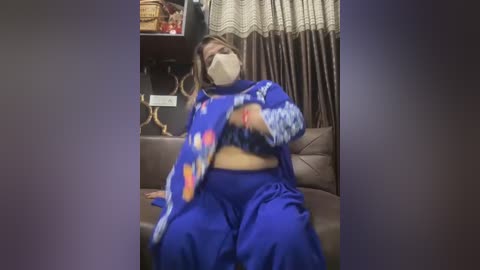 Media: Video of a plus-sized woman with fair skin, wearing a blue sari with floral patterns, seated on a brown couch, wearing a face mask, in a dimly lit room with brown curtains and a basket in the background.