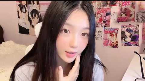 Media: Video of a young Asian woman with long black hair, fair skin, and light makeup, wearing a white top. Background features a bed with white sheets and a wall covered with Japanese magazines and posters.