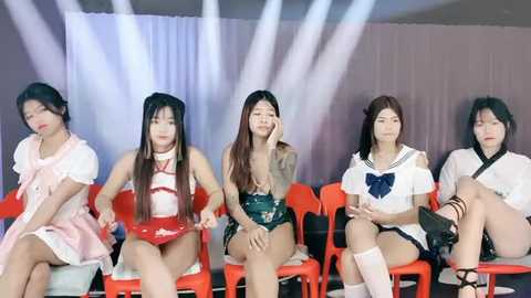 Media: Video of five Asian women in colorful, revealing costumes seated on bright orange chairs against a white and purple backdrop. They have various hairstyles and expressions, suggesting a theatrical or promotional setting.