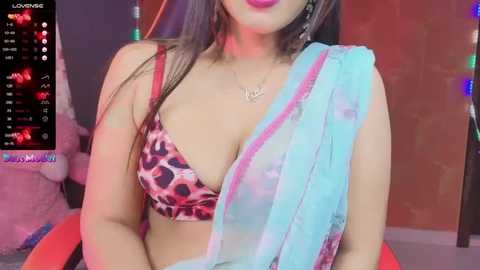 Media: Video of a South Asian woman with long black hair, wearing a leopard print bra and a see-through blue and pink sari, standing in a vibrant, colorful room.