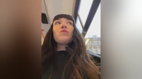 Media: Video of a young woman with long brown hair and bangs, wearing a black coat, looking out a bus window at an urban cityscape.