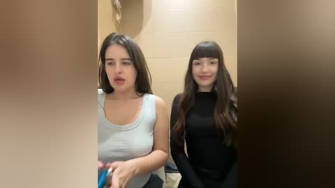 Media: Video of two young women in a restroom. The woman on the left has long dark hair, wears a white tank top, and appears surprised. The woman on the right has long dark hair, wears a black long-sleeve shirt, and smiles.