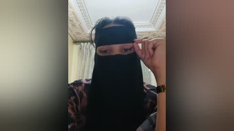 Media: A video of a woman in a black niqab, revealing only her eyes and part of her hand, standing in a lavish room with intricate ceiling moldings and white curtains.