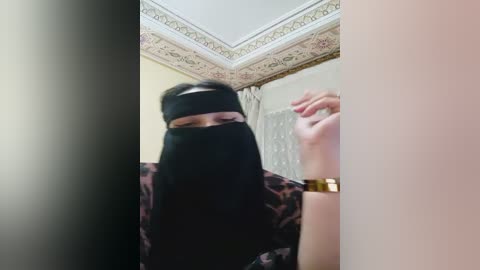 Media: Video of a person wearing a black niqab and gold watch, standing in a room with ornate ceiling molding and beige walls.