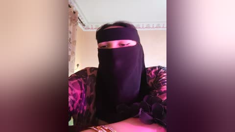 Media: Video of a person wearing a black niqab, sitting on a bed with leopard print sheets, in a dimly lit room with beige walls and a ceiling fan.