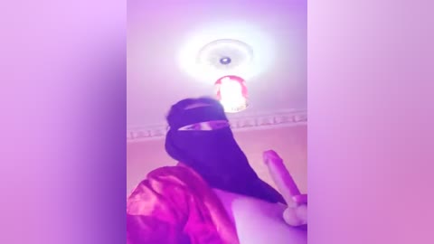 Media: Video of a person with black hair, wearing a black mask and a pink robe, kneeling in a bathroom with a bright, circular light above, holding a dildo.