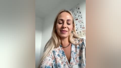 Media: Video of a smiling, fair-skinned blonde woman with long hair, wearing a floral blouse, standing in a room with white walls and a chalkboard scribbled with mathematical equations.