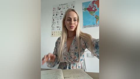 Media: Video of a blonde woman with medium build, wearing a floral dress, sitting at a desk with an open book, in a classroom with educational posters on the wall.